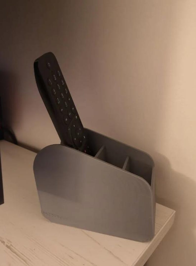 May include: A grey plastic remote control tidy with three compartments, holding a black remote. The tidy is on a white surface, with a light-coloured wall in the background. The holder is designed to organise remotes.