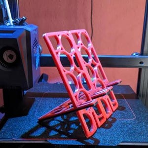 May include: A bright red, 3D-printed phone stand. The stand has a geometric design with circular cutouts and angled supports. It is sitting on a dark blue surface, with a 3D printer in the background.