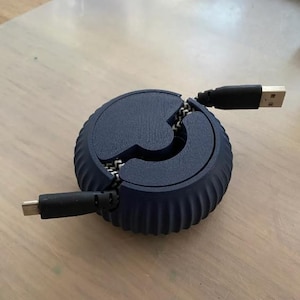 May include: A dark blue, circular cable tidy with a zip-like design. The organiser has a ribbed exterior and holds a USB cable and a USB-C cable. The cables extend from the sides, ready for use.