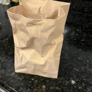 May include: A crumpled, open-top brown paper bag stands upright on a dark countertop. The bag's texture is visibly creased and folded, suggesting it has been used. The lighting highlights the bag's shape and the shadows within its folds.