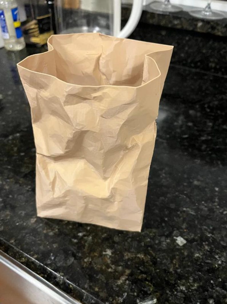 May include: A crumpled, open-top brown paper bag stands upright on a dark countertop. The bag's texture is visibly creased and folded, suggesting it has been used. The lighting highlights the bag's shape and the shadows within its folds.