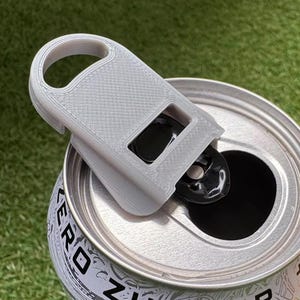 May include: A gray 3D-printed can opener attached to a silver can. The opener has a loop for easy handling and a rectangular opening to lift the tab. The can is open, revealing a black interior.