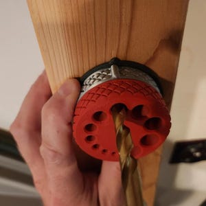 May include: A red drill guide with multiple holes is attached to a wooden piece. A drill bit is inserted through the guide. The guide is held in place by a black and silver ring.