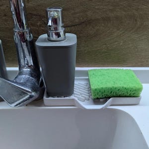 May include: A grey soap dispenser with a silver pump sits on a white tray next to a green sponge. A chrome tap is in the background. The items are on a white surface with a wood-grain background.