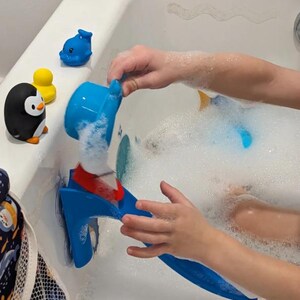 May include: A blue bath toy with a red accent is being used in a bathtub filled with bubbles. Rubber duckies, a penguin, and a dolphin bath toy are also visible. The scene suggests a fun and playful bath time experience for children.