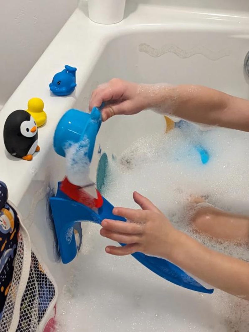 May include: A blue bath toy with a red accent is being used in a bathtub filled with bubbles. Rubber duckies, a penguin, and a dolphin bath toy are also visible. The scene suggests a fun and playful bath time experience for children.