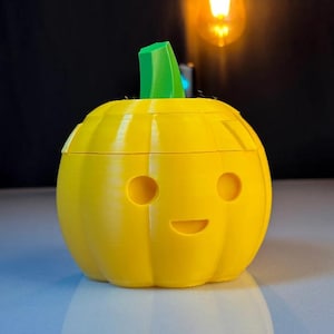 May include: A yellow 3D-printed pumpkin-shaped container with a green stem. The pumpkin has two circular eye cutouts and a curved mouth, giving it a friendly expression. The container is sitting on a reflective surface.
