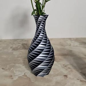 May include: A decorative vase with a geometric pattern of black, gray, and white triangles. The vase has a twisted design and holds green stems with white flowers. The vase is on a marble-like surface.