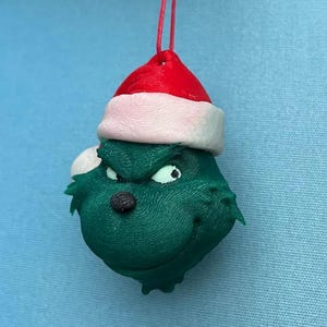 May include: A green Grinch-themed Christmas ornament with a Santa hat. The ornament has a red hat with a white brim, a black nose, and white eyes. The ornament is hanging from a red string.