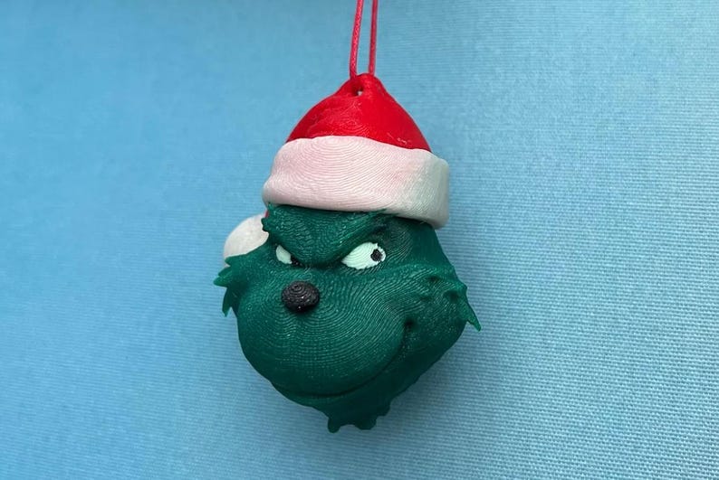 May include: A green Grinch-themed Christmas ornament with a Santa hat. The ornament has a red hat with a white brim, a black nose, and white eyes. The ornament is hanging from a red string.