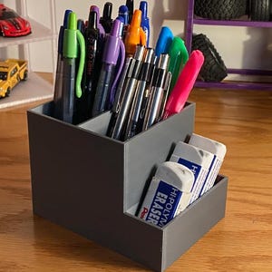 May include: A gray desk organizer with multiple compartments, holding pens, pencils, and erasers. The organizer is rectangular with a stepped design, providing easy access to stationery items. The erasers are white with blue text.