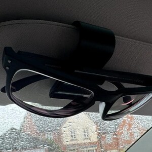 May include: Black eyeglasses clipped to a car visor. The glasses have a black frame with clear lenses. The visor is gray and the background shows a residential area with a rainy window.