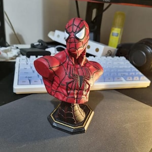 May include: A red and black Spider-Man bust figurine. The bust features the character's iconic mask with white eyes and a detailed web pattern. The figurine is mounted on a black and gold hexagonal base.
