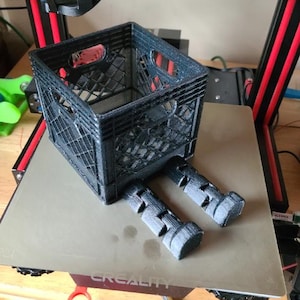 Puede incluir: Una pequeña caja de leche negra impresa en 3D y dos soportes a juego se encuentran en la plataforma de una impresora 3D. La impresora tiene un marco rojo y negro. La caja es una pequeña réplica detallada de una caja de leche estándar.