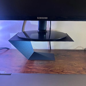 Puede incluir: Un soporte de monitor de metal negro con un diseño geométrico. El soporte sostiene un monitor Samsung negro. El soporte está sobre un escritorio de madera. Un portátil es visible en primer plano.
