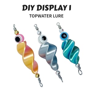 May include: Three topwater fishing lures with a spiral design in yellow-pink, silver, and teal. Each lure has a large eye and a metal hook. The text "DIY DISPLAY I TOPWATER LURE" is at the top.
