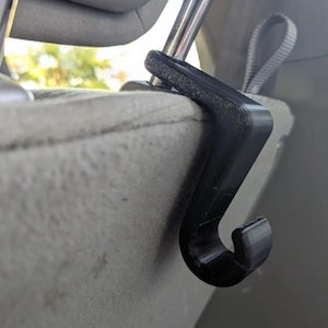 May include: A black plastic car seat headrest hook. The hook is designed to attach to the headrest posts and provides a convenient place to hang items. The hook is curved and has a smooth finish.