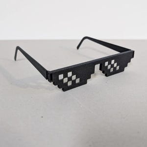 May include: Black pixelated sunglasses with a rectangular frame and square lenses. The sunglasses have a classic design, with a bridge and arms. The glasses are made of a matte material and sit on a light gray surface.