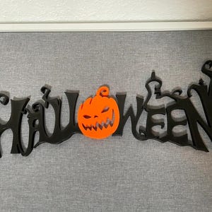 Może przedstawiać: Czarny znak Halloween ze słowem "Halloween" i wyrzeźbioną pomarańczową dynią pośrodku. Znak jest wykonany z ciemnego materiału i jest zamontowany na szarej powierzchni.
