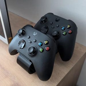 May include: Two black Xbox controllers are displayed. The controllers have the standard button layout with colored A, B, X, and Y buttons. One controller is resting on a black stand.