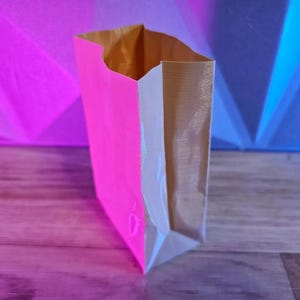 May include: A paper bag with a neon pink front panel and a natural brown interior. The bag is open at the top, revealing the interior. The background is a gradient of pink, blue, and purple.