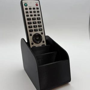 Puede incluir: Un soporte negro para mando a distancia con tres compartimentos, que contiene un mando a distancia plateado y negro. El mando tiene muchos botones y el texto "POWER ON" y "SET". El soporte está hecho de un material texturizado.