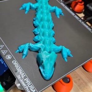 May include: A turquoise 3D-printed lizard figurine with detailed scales and limbs, resting on a black surface. The figurine is the focal point, showcasing the intricate design and vibrant color. The base has the text "ORIGINAL PRUSA".