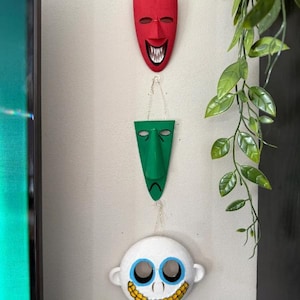 Pode incluir: Três máscaras decorativas penduradas em uma parede. A máscara superior é vermelha com um rosto sorridente, a máscara do meio é verde com um rosto triste e a máscara inferior é branca com um rosto sorridente. As máscaras são conectadas por correntes.