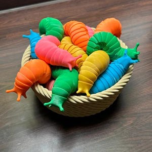 May include: A woven basket filled with colorful, segmented slug toys in shades of green, orange, yellow, pink, and blue. The toys have antennae and are arranged on a dark wooden surface.