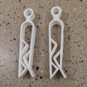 Puede incluir: Dos pinzas de plástico blanco con un diseño único. Cada pinza tiene una parte superior circular, un cuerpo curvo y un patrón geométrico en el centro. Las pinzas están diseñadas para sujetar objetos.