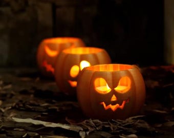 May include: Three lit pumpkin candle holders with carved faces, arranged in a row on a surface with dried leaves. A fourth, out-of-focus pumpkin is in the background. The pumpkins are orange and have a warm glow.