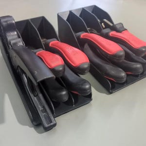 Puede incluir: Dos organizadores de herramientas negros que contienen herramientas con mango negro y rojo. Los organizadores tienen múltiples ranuras para sujetar las herramientas. Las herramientas parecen ser alicates y otras herramientas manuales.