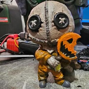 May include: A handcrafted figurine of Sam from the horror film Trick 'r Treat. The figure is wearing an orange jumpsuit and holding an orange pumpkin-shaped lollipop. The head is made of burlap with button eyes. The figurine is approximately 5 inches tall.