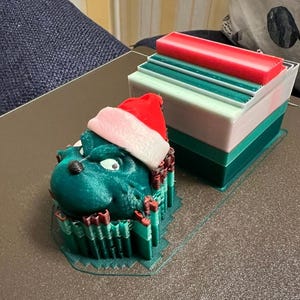 May include: A 3D-printed Christmas decoration featuring a green Grinch-like character wearing a Santa hat. Beside it is a layered block with red, white, and green sections. The objects are on a dark surface, suggesting a festive theme.