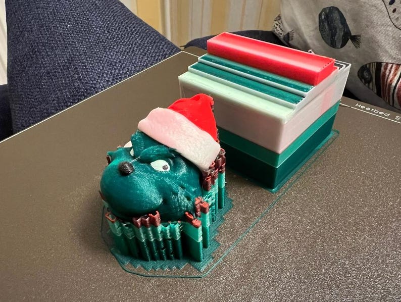 May include: A 3D-printed Christmas decoration featuring a green Grinch-like character wearing a Santa hat. Beside it is a layered block with red, white, and green sections. The objects are on a dark surface, suggesting a festive theme.