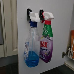 May include: Two spray bottles hanging on a white cabinet door. One bottle is clear with blue liquid and a white and blue sprayer. The other is purple and green with the text "VAX Spray & Wipe".