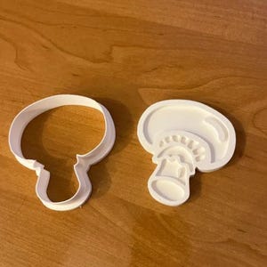 May include: Two white plastic mushroom-shaped cookie cutters. One is a simple outline of a mushroom, and the other is a detailed cross-section. The cutters are on a wooden surface.