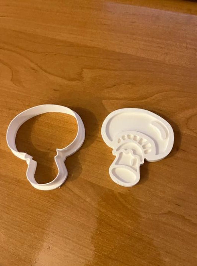 May include: Two white plastic mushroom-shaped cookie cutters. One is a simple outline of a mushroom, and the other is a detailed cross-section. The cutters are on a wooden surface.