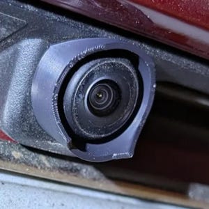 Puede incluir: Primer plano de una lente de cámara de coche negra con una carcasa de plástico gris oscuro. La cámara está montada sobre una superficie roja, con una parte de la carrocería del coche visible. La lente está centrada dentro de la carcasa.