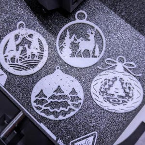 May include: Four white Christmas ornaments with intricate cut-out designs. Each ornament features a different winter scene: deer in a forest, a deer with trees, a snowy forest, and a Christmas tree with a bow.