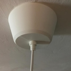 Puede incluir: Un plafón blanco con una pantalla cónica y una base circular. La lámpara está suspendida del techo por un cable blanco. La lámpara está hecha de un material liso y mate.