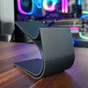 May include: A black, curved stand, likely 3D-printed, with a textured surface. The stand has a base and a curved top section, designed to hold a phone or tablet. The stand is on a wooden surface.