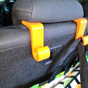 May include: Bright orange car seat headrest hooks. The hooks are made of plastic and are designed to hang bags or other items. The hooks are attached to the headrest of a car seat. A patterned tote bag is hanging from one of the hooks.