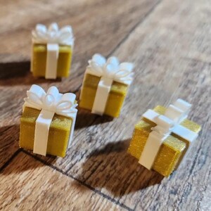 May include: Four miniature gift boxes with white bows and ribbons. The boxes are gold with a glittery texture, and the ribbons are white. The gifts are arranged on a wooden surface.