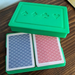 May include: A green plastic playing card case with a lid featuring embossed suit symbols. Inside, two decks of cards are visible, one with a blue patterned back and the other with a red patterned back. The case sits on a wooden surface.