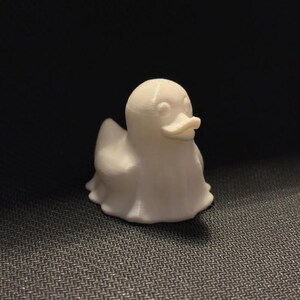 May include: A small, white, 3D-printed duck figurine. The duck has a rounded body, a small beak, and a ghost-like appearance. The figurine is sitting on a dark, textured surface.