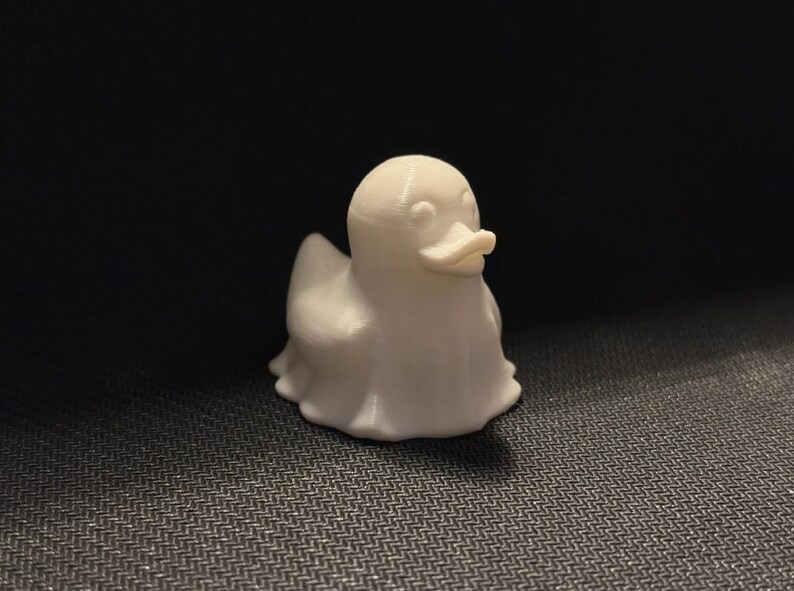 May include: A small, white, 3D-printed duck figurine. The duck has a rounded body, a small beak, and a ghost-like appearance. The figurine is sitting on a dark, textured surface.