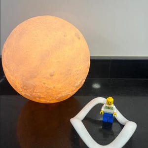 May include: A glowing, spherical moon lamp with a textured surface, emitting a warm, orange light. A white, curved, geometric shape and a Lego figure with a yellow head, white shirt, and blue pants are in the foreground.