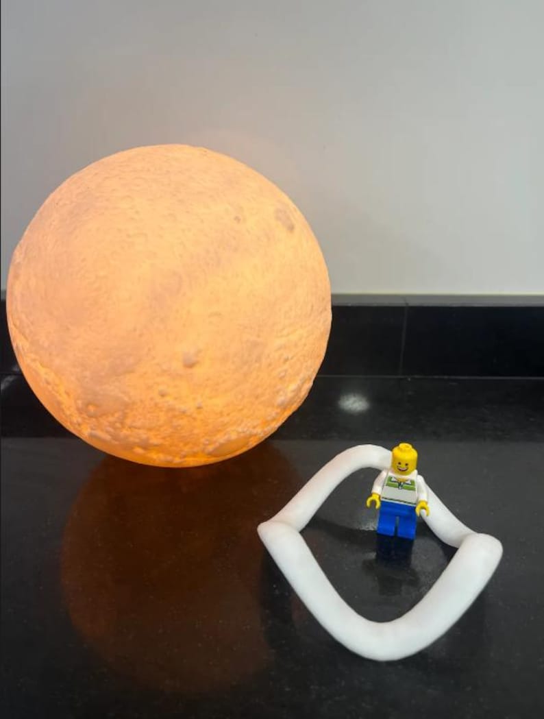 May include: A glowing, spherical moon lamp with a textured surface, emitting a warm, orange light. A white, curved, geometric shape and a Lego figure with a yellow head, white shirt, and blue pants are in the foreground.