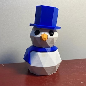 May include: A geometric snowman figurine with a blue top hat, scarf, and eyes. The snowman has a white body, a blue scarf, and an orange nose. The figure is 3D printed and sits on a red surface.